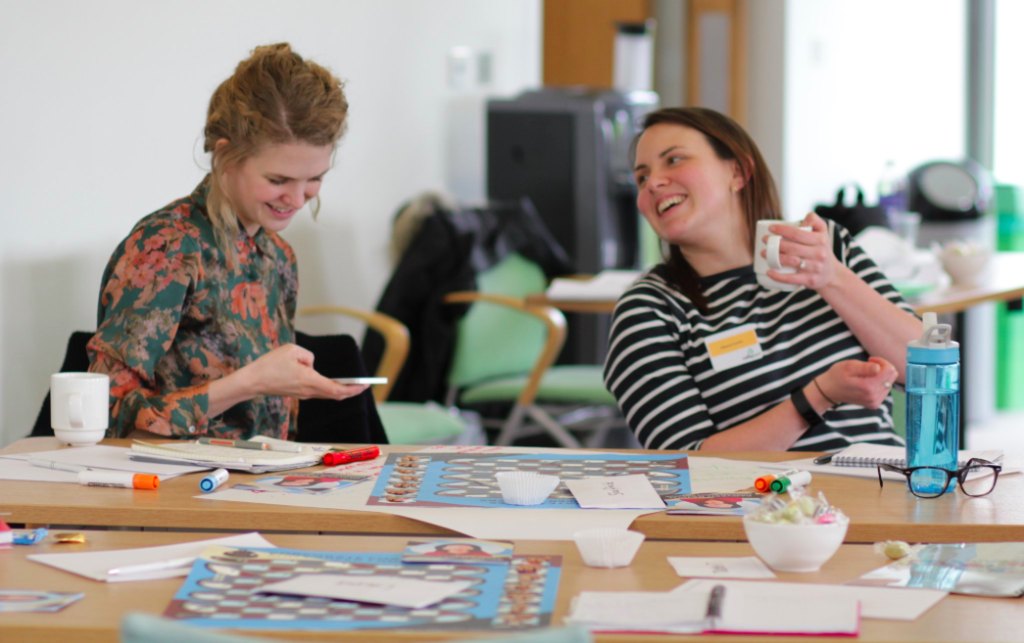 Two participants laughing at a table during the Corporate Snakes and Career Ladders simulation, with game boards and markers in front of them