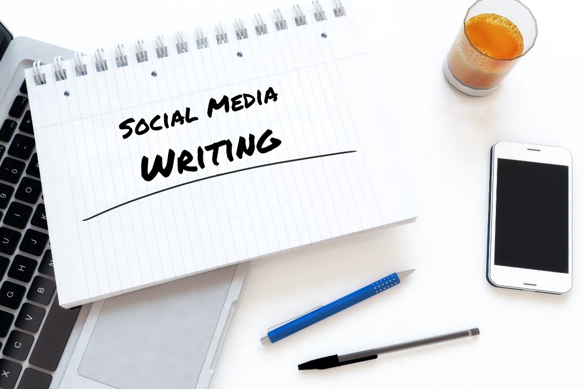 Notebook with "Social Media Writing" handwritten on it, beside a laptop, smartphone and pens on a white desk